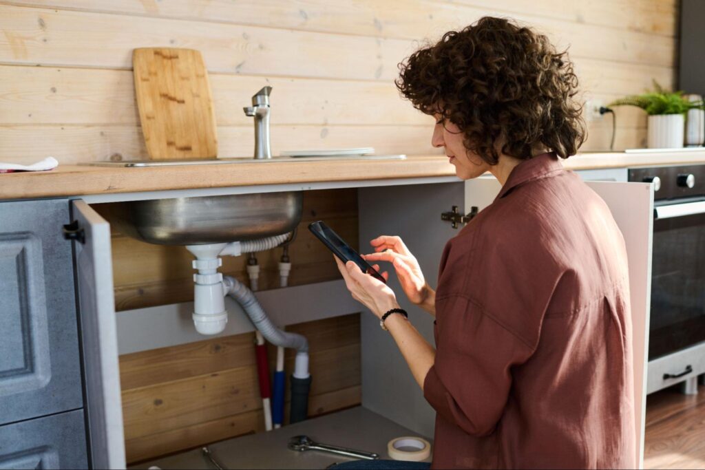 Checking kitchen plumbing while using a smartphone for maintenance tips.