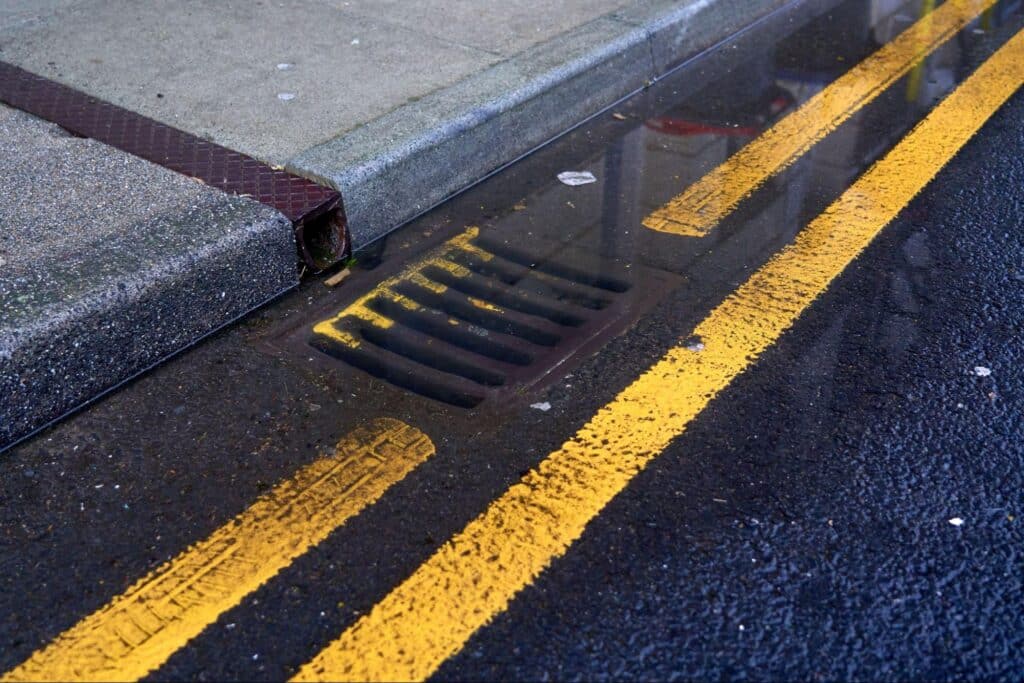 Wet road with yellow lines and a drain reflecting puddles after rain.