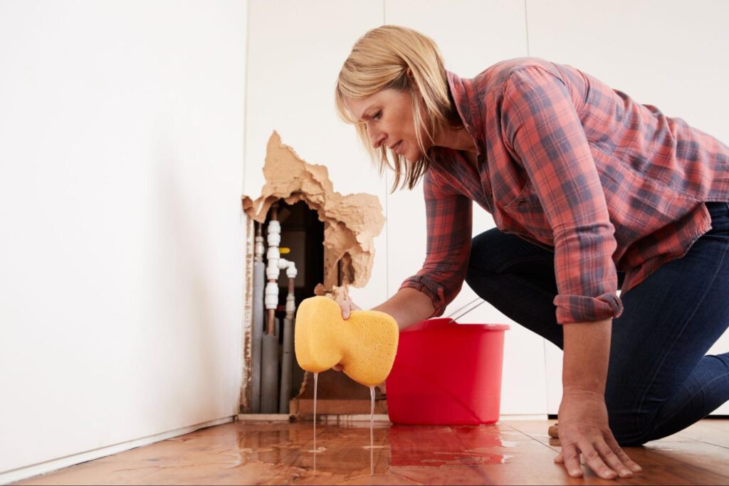 Woman cleaning water damage from a burst pipe with sponge and bucket in home renovation project.