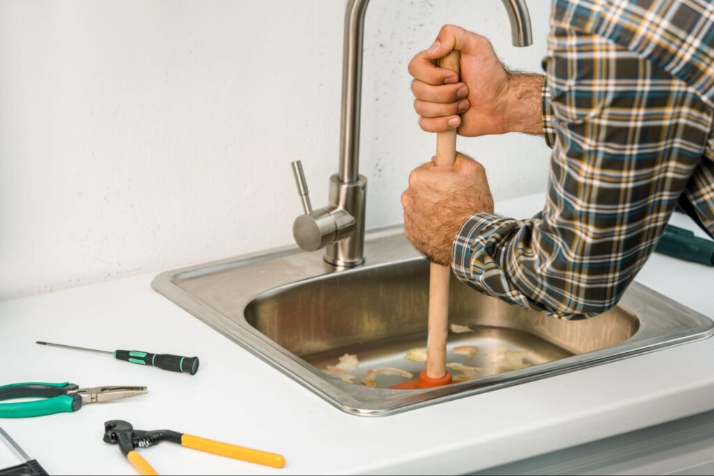 Man using a plunger to unclog a kitchen sink with plumbing tools nearby.