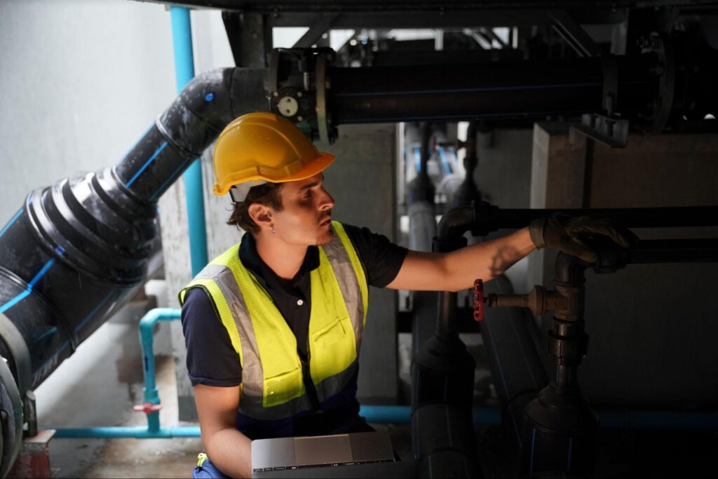 Worker in safety gear inspecting industrial machinery and equipment for routine plumbing maintenance.
