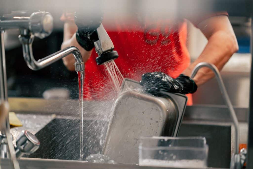 Hands washing a metal pan under running water in a commercial kitchen.