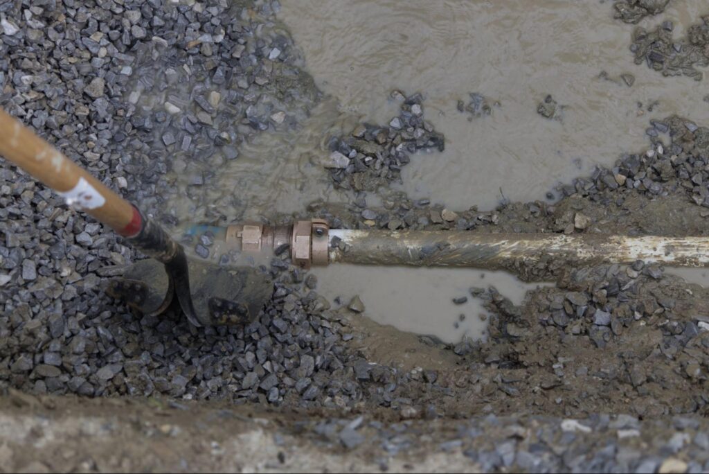 Excavation for plumbing revealing a water pipe, surrounded by gravel and muddy soil.