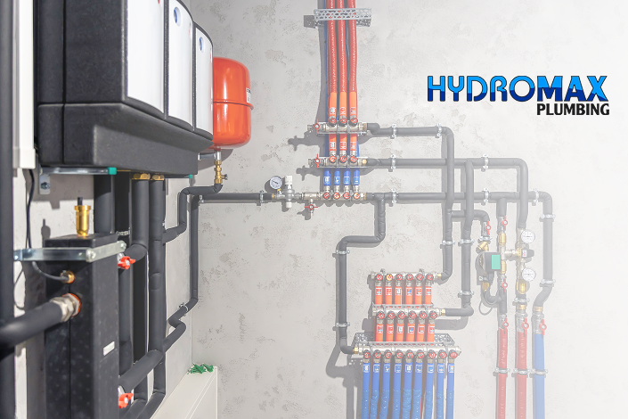 Modern plumbing system with black pipes and red valves, arranged neatly on a light gray wall. The logo "Hydromax Plumbing" is visible. The setting is organized and technical.