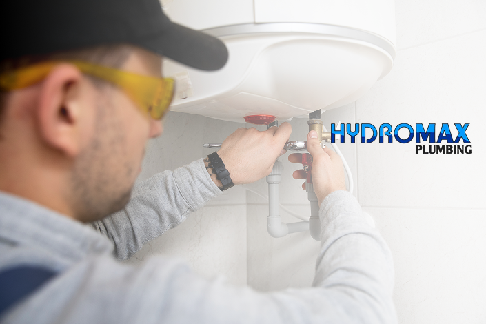A plumber in a cap and safety goggles installs a water heater with tools on a wall. The "Hydromax Plumbing" logo is prominently displayed beside him.