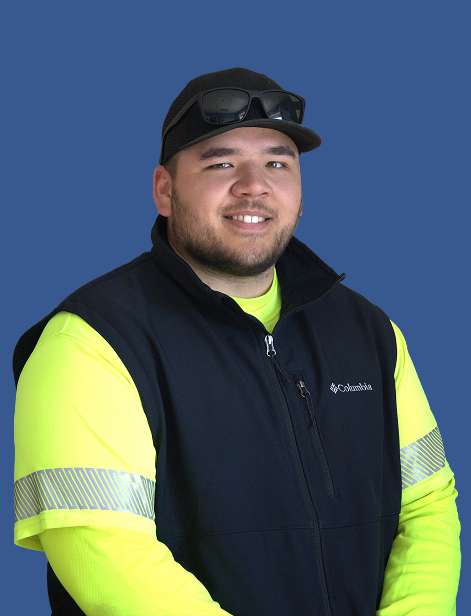 A man in a casual outdoor outfit stands against a blue background. He wears sunglasses on a cap, a black vest over a neon yellow shirt, and smiles warmly at the camera.