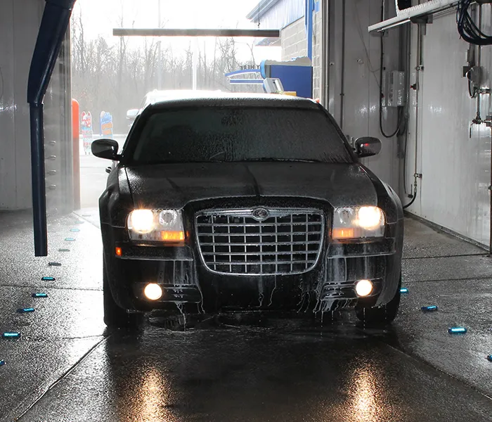A black car with headlights on is covered in water and foam as it exits a dimly lit car wash. The scene is wet, with reflections on the ground.