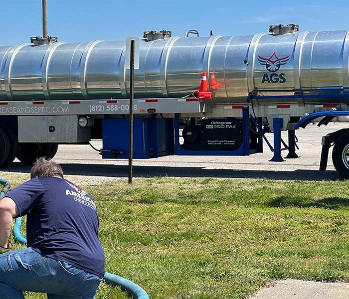 A man in a blue shirt works with a hose near a shiny silver tanker truck labeled "AGS" on a sunny day. The scene is industrious and focused.