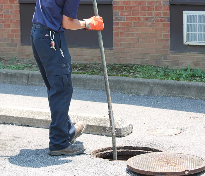 A worker in blue uniform uses a pole to inspect a manhole. The person wears orange gloves and stands on a paved street next to a raised brick building.