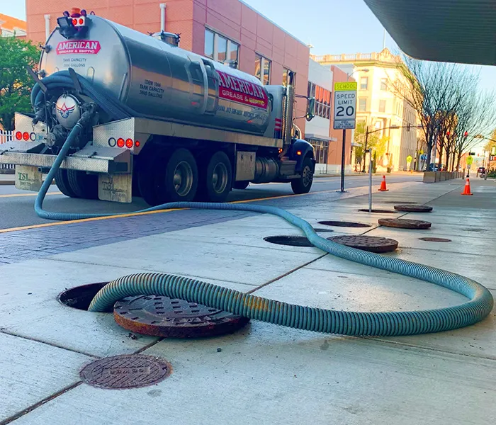 A large truck labeled "American" pumps waste through a long hose into open manholes on a city street. Speed limit sign and traffic cones visible.