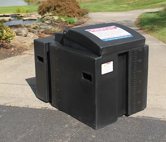 Large black plastic weight container on a paved path, with ruler markings and safety labels, set against a background of grass and rocks near a pond.