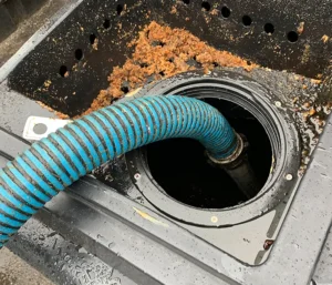 A blue and black striped hose is inserted into a circular opening of a wet, dirty container with brown residue on the sides, suggesting cleaning or drainage.