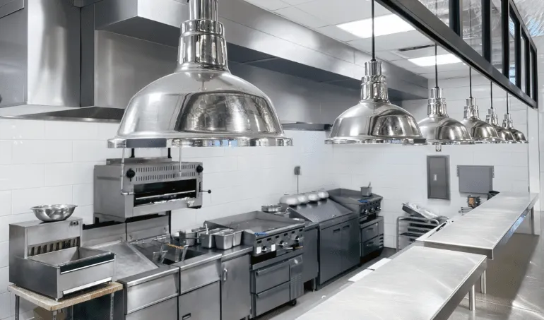 Sleek industrial kitchen with stainless steel appliances, white tile walls, and large metallic pendant lights. The space is modern and immaculate.